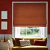 Burnt-orange Roman blind, lowered to the sill with soft folds; blocking light and framing a white-paned window; living room with mustard sofa, cushions, red walls, framed sketches and garden view.