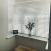 White horizontal slatted blinds covering a bay window, mostly closed and filtering soft daylight; on the windowsill a glass vase with green stems, framed photo, and sheepskin seat below.