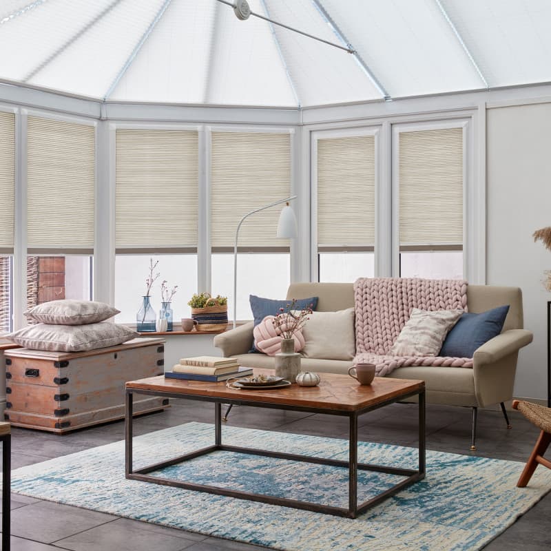 Beige pleated cellular blinds across tall conservatory windows, lowered and filtering soft daylight; a cozy sitting area with beige sofa, chunky pink throw, wooden coffee table and blue-pattern rug.
