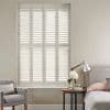 White plantation shutters—four tall louvered panels—fully closed, softly filtering daylight into a neutral bedroom corner with upholstered headboard, bedside table, lamp, vase of flowers and a gray armchair.