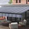 Striped retractable fabric awning with blue-gray diagonal stripes, fully extended over a patio; shading glass doors and outdoor seating — daytime, brick upper facade, wooden deck, lounge chairs and coffee table.