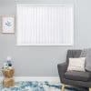 White vertical blinds covering a window, fully closed with scalloped bottom tabs, filtering daylight; in a gray-walled living room with gray armchair, knitted throw, wooden side table and blue rug.