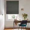 Object: dark grey Roman blind lowered over a window, fabric with a small gap at bottom. Action: partially closed, filtering daylight. Context: home desk vignette with chair, mirror, vase of flowers.