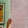 Pink fabric blind with white leaf pattern, held at its left edge by a hand; pale green interior wall with framed colorful artwork, softly lit.