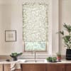 Patterned roller blind covers window, filtering light over a kitchen sink; stainless faucet, wooden cabinets, countertop with bowl of greens, potted plants and framed sketch on wall.