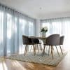 Object: sheer floor-to-ceiling pleated curtains on two walls, pale gray with vertical folds. Action: drawn closed, softly filtering daylight. Context: sunlit dining nook with round table, four chairs, patterned rug.