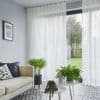 Sheer floor-to-ceiling pleated curtains, partially drawn, softly filter daylight through sliding glass doors into a living room with a pale sofa, patterned tile floor, potted plants, and a lawn view.