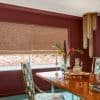 Beige woven Roman shades, fully lowered with visible horizontal folds, filtering soft daylight through the window; burgundy dining room with a wooden table, floral vase, patterned chairs, brass chandelier.