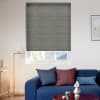 Gray pleated cellular blind lowered over a recessed window, blocking light with a narrow gap at bottom; modern living room context with blue sofa, colored cushions, floor lamp, and daylight.
