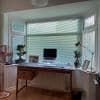 Horizontal pale green Venetian blinds covering a three-panel bay window, mostly closed, filtering soft daylight into a tidy home office with wooden desk, laptop, adjustable lamp, and potted plants.