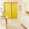 Two tall yellow pleated blinds covering side-by-side windows, fully lowered and filtering sunlight; modern bright kitchen-dining area with white furniture, wooden floor, table, vase and fruit.