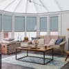 Pale blue cellular blinds covering conservatory windows, lowered and gently filtering daylight; above a cozy sunroom with a beige sofa, pink chunky knit throw, wooden coffee table and blue rug.