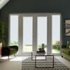 Glass French doors with textured white roller blinds cover most panes, framing a sunlit garden; a glass coffee table rests on a chevron rug between green armchairs in living room.