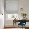 Pale blue wavy-patterned Roman blind covers most of the window, partially lowered and filtering daylight; desk with chair, mirror and vase of flowers sits beneath in a lit study.
