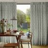 Leaf-patterned pleated curtains, parted to reveal a sliding glass door; filtering soft daylight into a breakfast nook with wooden table, chairs, pendant lamp, and a green garden view.