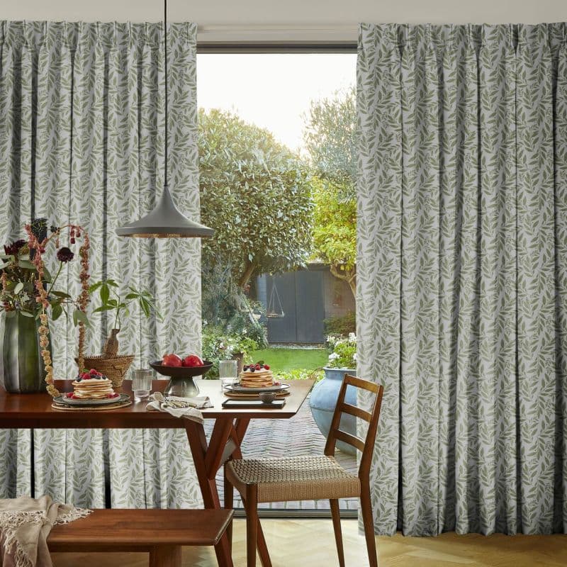 Leaf-patterned pleated curtains, parted to reveal a sliding glass door; filtering soft daylight into a breakfast nook with wooden table, chairs, pendant lamp, and a green garden view.