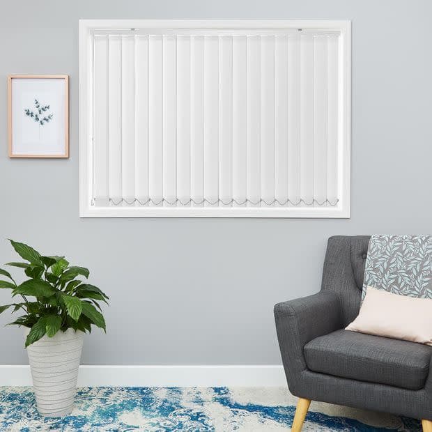 White vertical blinds covering a central window, closed and softly diffusing daylight; minimalist living room with pale grey wall, potted plant, framed botanical print, grey armchair and patterned rug.