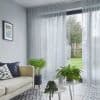 Sheer pale-gray floor-length curtains hung from ceiling track, softly pleated and partially drawn, filtering daylight; living room with beige sofa, patterned tiled floor, nesting white tables, potted plants, garden view.