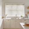 White horizontal Venetian blinds covering a wide window; slats closed, softly filtering daylight; positioned above a double stainless sink on a marble countertop in a bright, neutral kitchen with cream cabinetry and plants.