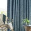 Textured teal curtains, floor-to-ceiling and mostly closed with a subtle crosshatch weave, filtering daylight; a wicker chair draped with a throw and a wooden side table holding a potted plant.