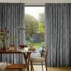 Full-length grey textured curtains with vertical striations, parted at center; framing and filtering sunlight onto a wooden dining table with pancakes and fruit, lamp and chairs, opening onto a garden.