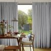 Object: light-gray, floor-to-ceiling pinch-pleat curtains parting at center; Action: framing a bright opening and admitting diffuse daylight; Context: wooden dining table set for breakfast, pendant lamp, garden view.