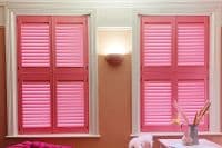 Bright pink louvered shutters, four-panel plantation style covering two tall side-by-side windows, fully closed and softly filtering pink light, in a cozy peach-walled room with a sconce and decorative vase.