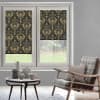 Patterned roller blinds (gold damask on black) lowered over a two-pane window, partially blocking light; modern living room with gray walls, rattan armchair, small table and backyard lawn view.
