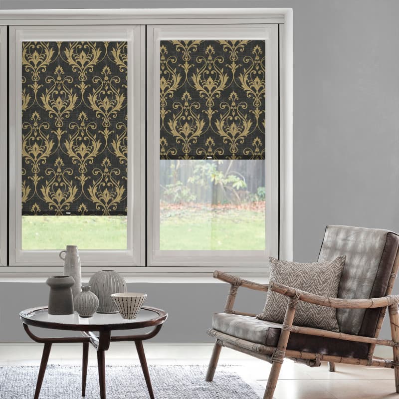 Patterned roller blinds (gold damask on black) lowered over a two-pane window, partially blocking light; modern living room with gray walls, rattan armchair, small table and backyard lawn view.