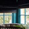 Dark velvet Roman blinds with fringed trim, partially lowered and dimming daylight, over turquoise-painted mullioned corner windows in a room with patterned seating, a plant; brick and garden visible outside.