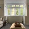 White slatted horizontal blinds covering a three‑pane bay window with decorative transom panels, partially open and filtering warm sunlight and leafy garden view into a neutral-toned living room with ottoman.