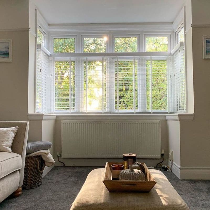 White slatted horizontal blinds covering a three‑pane bay window with decorative transom panels, partially open and filtering warm sunlight and leafy garden view into a neutral-toned living room with ottoman.