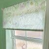 Floral roller blind with pastel green line-drawn flowers, lowered to mid-window, filtering bright daylight; mounted in a pale green-walled room over a frosted glazed sash window, metal pull chain visible.