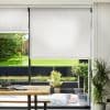 White roller blind, lowered mid-window with slight translucence, filtering daylight; overlooks sunlit backyard with lawn and potted plants, dining table and bench in the foreground.