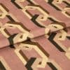 Geometric-patterned fabric with interlocking beige, brown and black hexagonal chains on a dusty rose background, folded across the center, lying on a flat surface under soft, neutral indoor lighting.