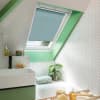 Light-blue pleated skylight blind, lowered halfway and filtering soft daylight; in a sloped attic bathroom with green-painted trim, white herringbone tiles, a basin, bathtub, and a potted plant.