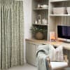 Pleated sage-green floral curtains, full-length and drawn closed, gently filtering daylight; beside a neutral built-in desk with wicker vase, iMac, lit candle, basket shelves, and a chair with throw.
Text: WHITE