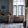 Beige floor-length curtains frame a tall window; white plantation shutters cover the lower sash with tilted horizontal slats, partially open and filtering daylight into a cozy study—desk and patterned wallpaper.