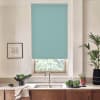 Teal roller blind covers a window above a kitchen sink; sunlight filters beneath it. Countertop holds bowls of greens, potted plants, faucet, towel and framed sketch.
