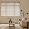 White horizontal blinds covering a large window, fully closed and softly diffusing daylight; neutral living room with beige sofa, wooden coffee table, sculptural vase, black floor lamp, patterned rug.