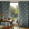 Floral blue pleated curtains with yellow blossoms, partly drawn framing a sliding glass door, letting daylight and a garden view into a dining area with table and wooden chairs.