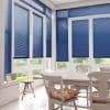 Blue horizontal Venetian blinds covering tall white-framed windows and doors, mostly lowered and filtering daylight; bright sunlit dining nook with a white table, chairs, floral cushion, stool and garden view.
