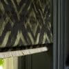 A patterned fabric Roman shade with olive-and-black geometric/ikat design and pale fringe trim hangs partially lowered and folded, covering a window within dark green trim, softly lit from the left.