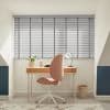 Grey horizontal Venetian blinds covering a wide skylight window, closed with slats angled to filter sunlight into an attic office; wooden desk, pink swivel chair, lamp, plant, soft daylight.