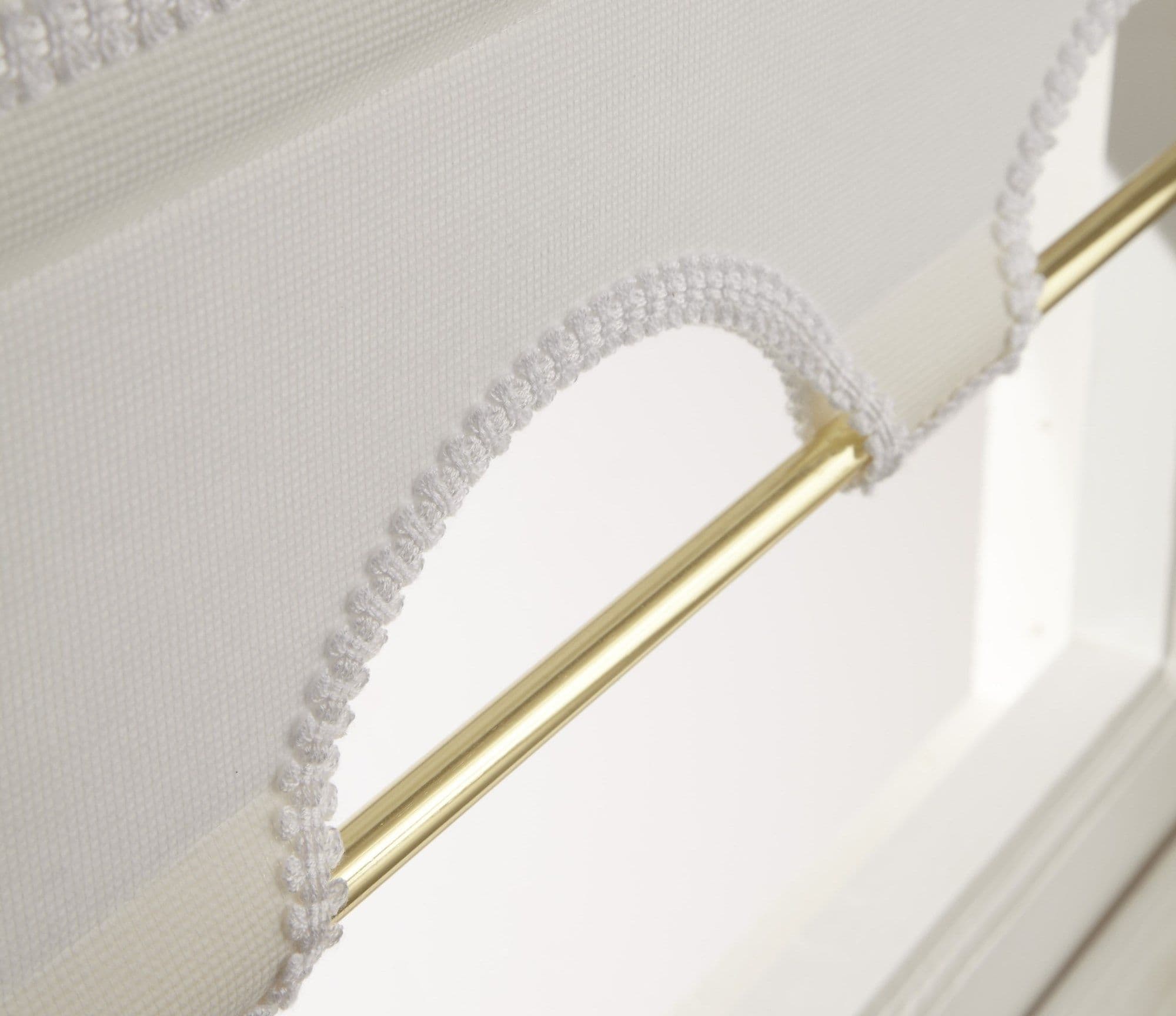 White scalloped‑trim fabric blind looped over a polished brass rod, crocheted edging forming decorative arches; partially raised, filtering soft daylight above white window frame in a bright room.