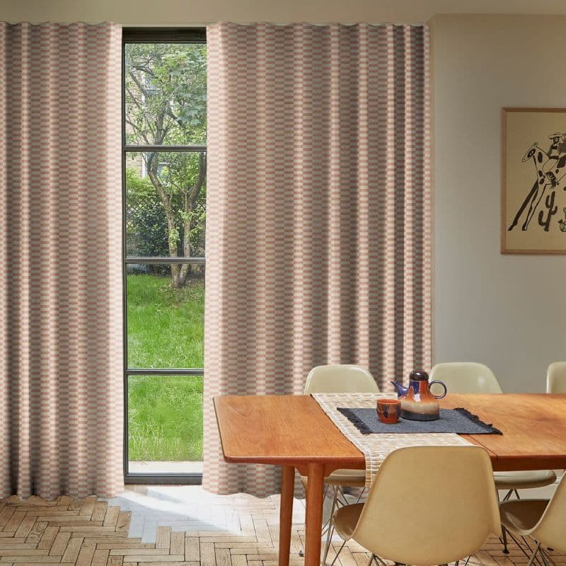 Blush-and-cream checkered full-length curtains, mostly drawn with a narrow central gap; allowing light to frame a mullioned window and green garden, in a sunlit dining room with wooden table.