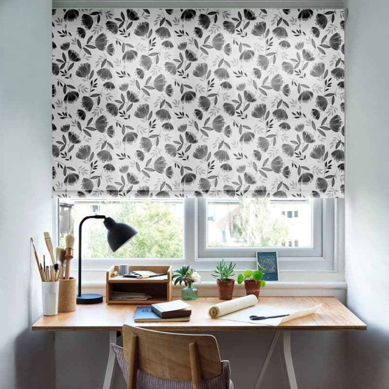 Floral roller blind with gray tulip-like pattern, lowered and filtering daylight; positioned over a double window above a small wooden desk with lamp, art supplies, potted succulents and chair.