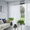 Sheer white floor-to-ceiling curtains on a ceiling track, softly filtering daylight and parted at a sliding glass door — living room with sofa, patterned tile floor, potted plants, garden view.