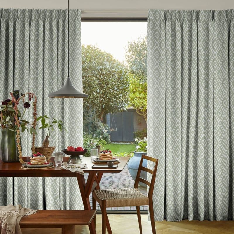Full-length pleated curtains with pale sage-grey diamond pattern, parted to reveal a sliding glass door, filtering morning light into a dining nook with wooden table, hanging lamp, and garden view.