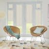 Beige roller blinds fitted inside white French doors and sidelights, lowered to partially filter daylight; bright living room with two wicker chairs, pastel throws, patterned rug and lawn view.